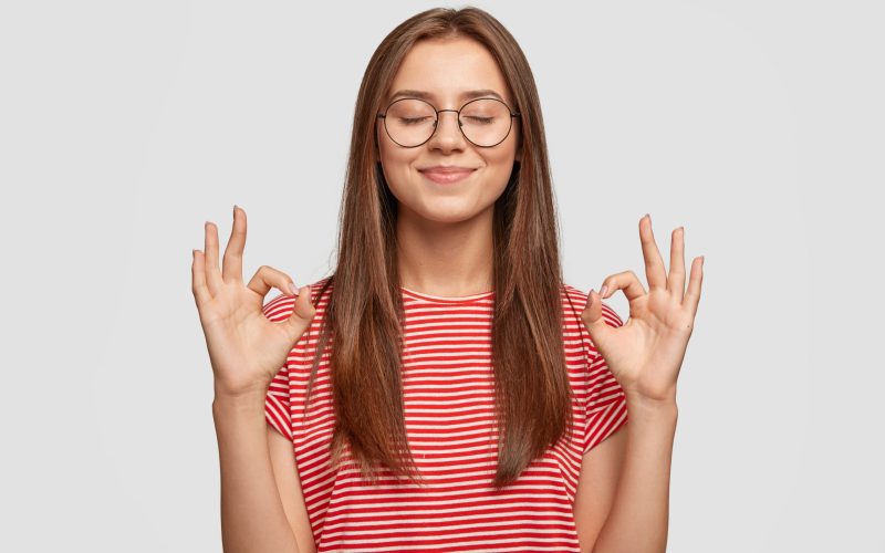 Pleased satisfied young female model makes zero gesture, wears transparent glasses, has long dark hair, dressed in casual striped t shirt, stands over white background. People, body language concept